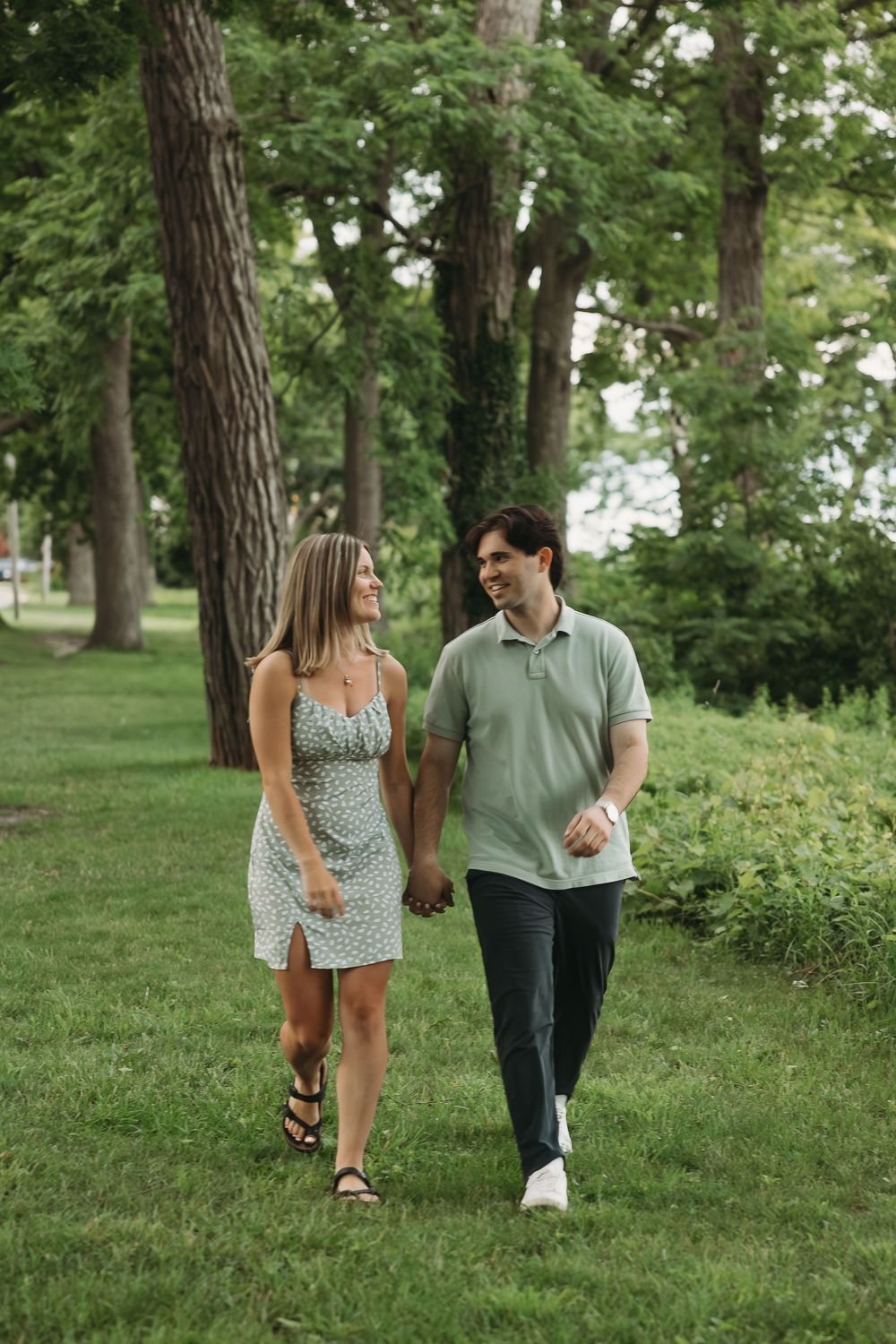A smiling couple walks hand-in-hand through a lush green park, she in a floral dress, he in a sage polo shirt.