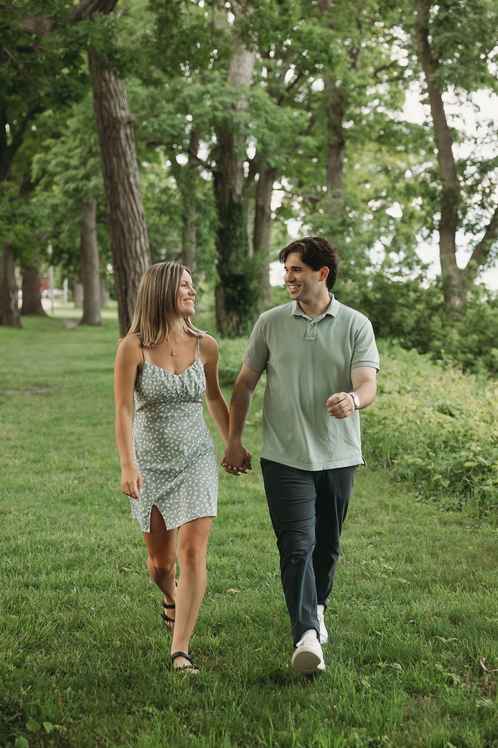 A smiling couple holds hands while walking through a lush green park, woman in floral dress, man in sage polo shirt.