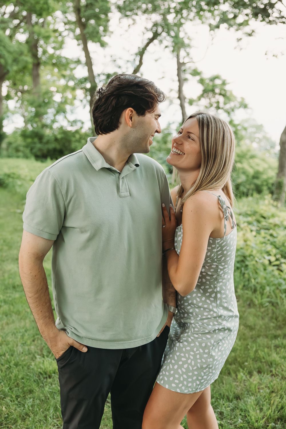 A smiling couple in matching sage green outfits share a joyful moment together in a lush green park.