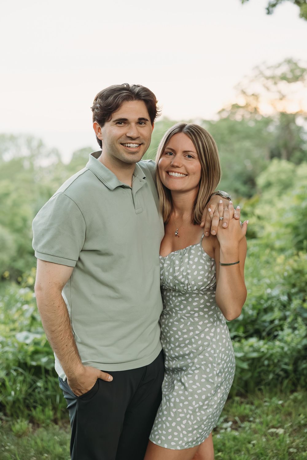Happy couple posing outdoors in matching sage green outfits, woman displaying engagement ring, lush greenery behind.