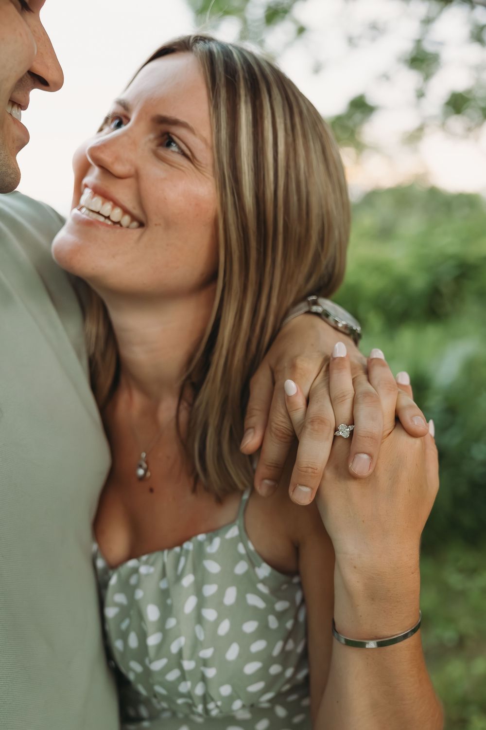 Smiling woman with engagement ring holds hands with man outdoors, wearing a sage green floral dress in a romantic couples photo.