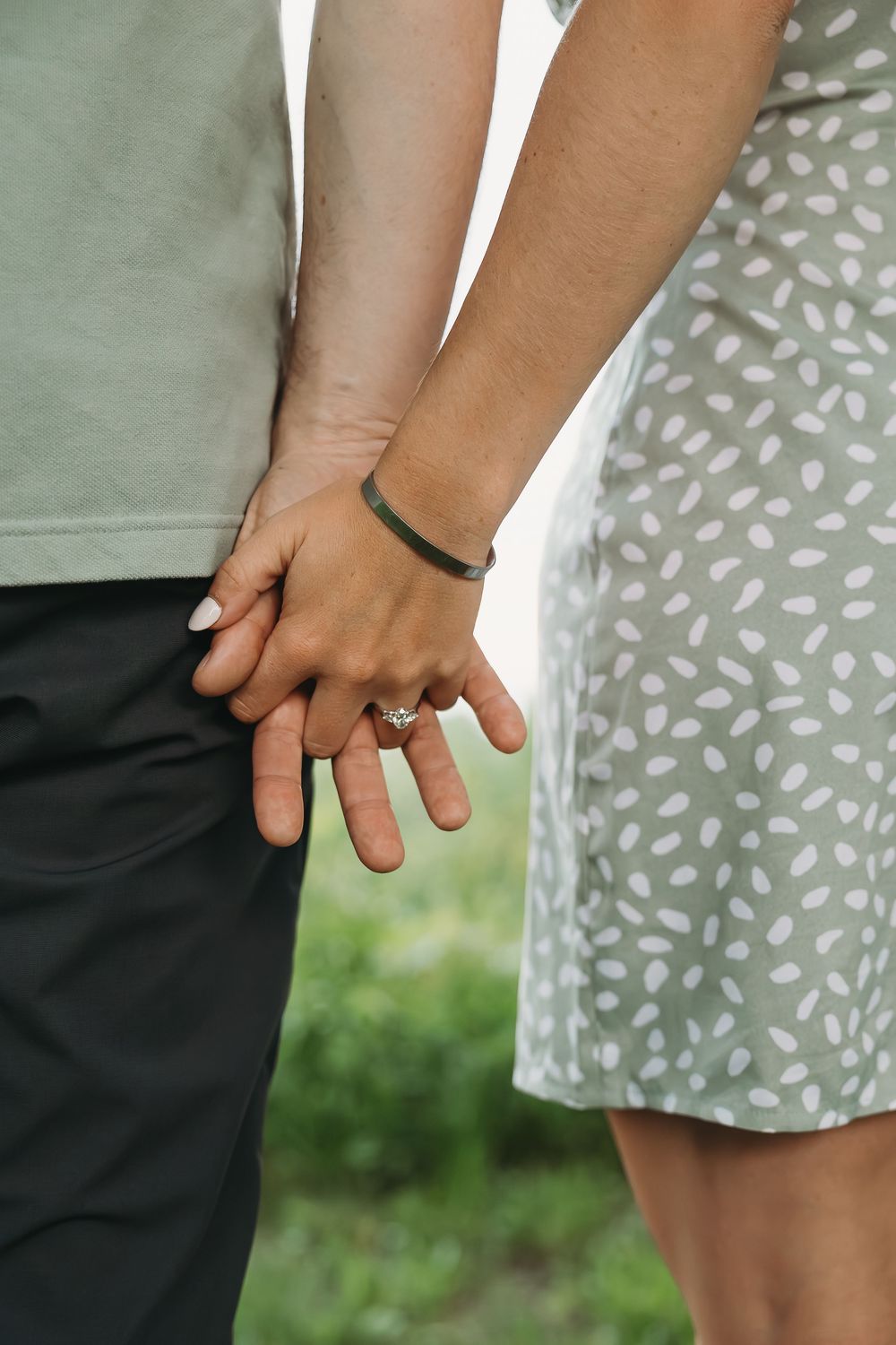 Close-up of couple holding hands, woman wearing diamond engagement ring and green bracelet, man in black pants.