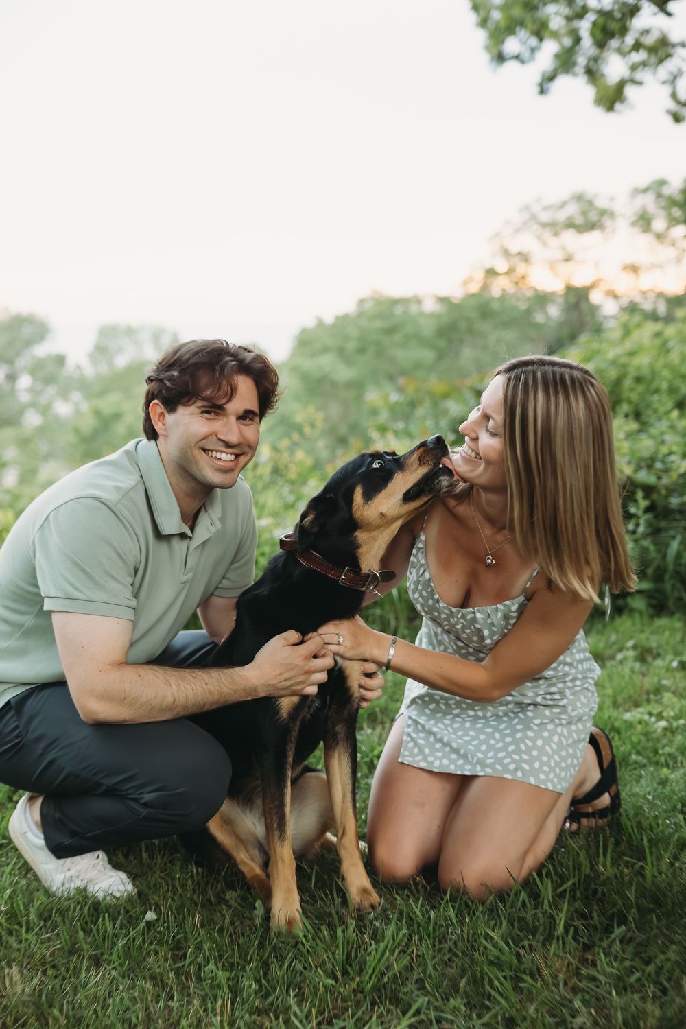 A smiling couple crouches on green grass with their black and tan dog licking the woman's face outdoors.