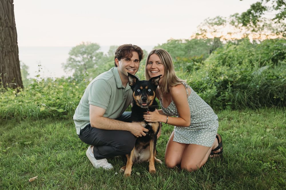 Couple smiling with black and tan dog outdoors on green grass near trees during golden hour.