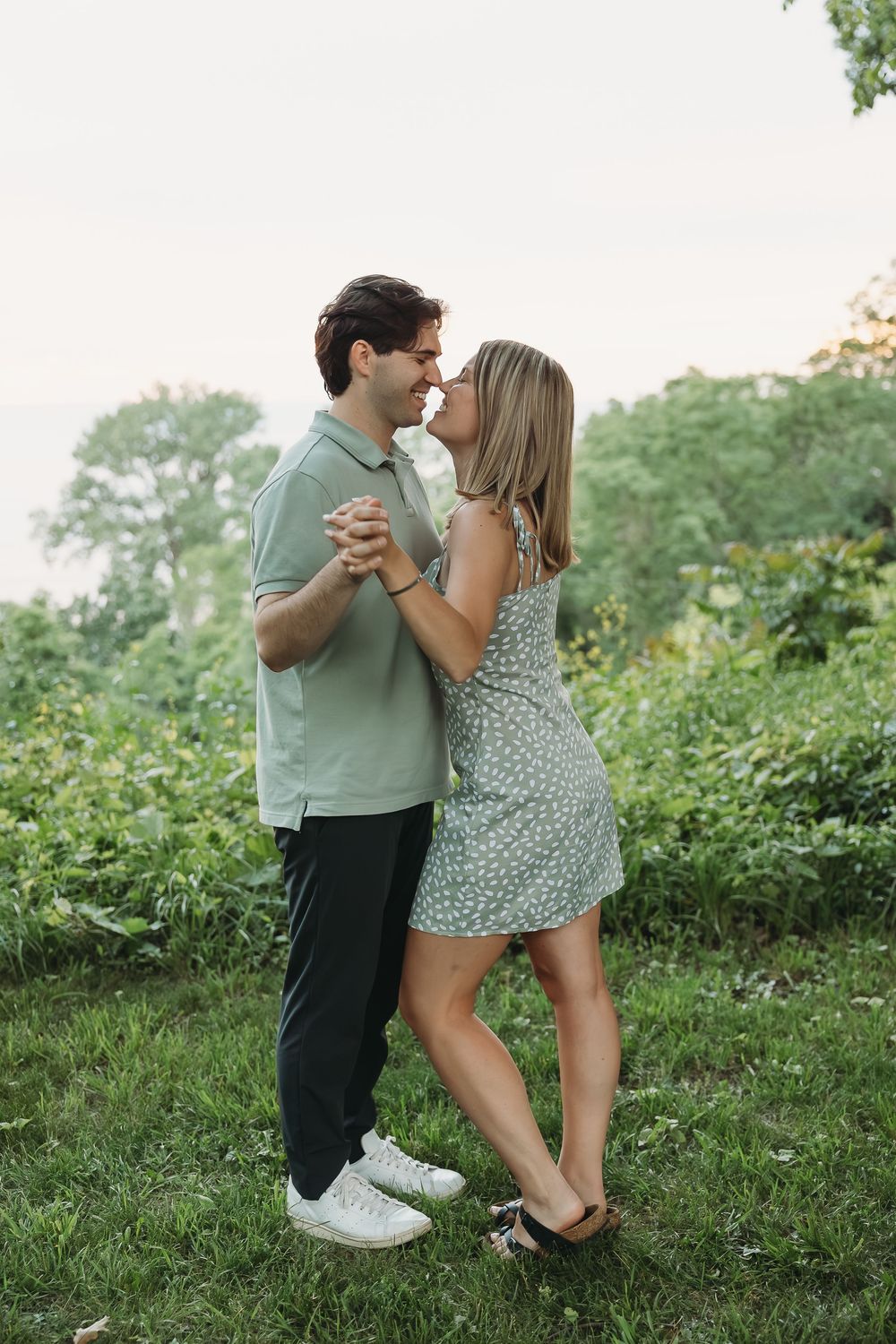 A couple dances closely outdoors, wearing matching sage green outfits, surrounded by lush greenery at sunset.