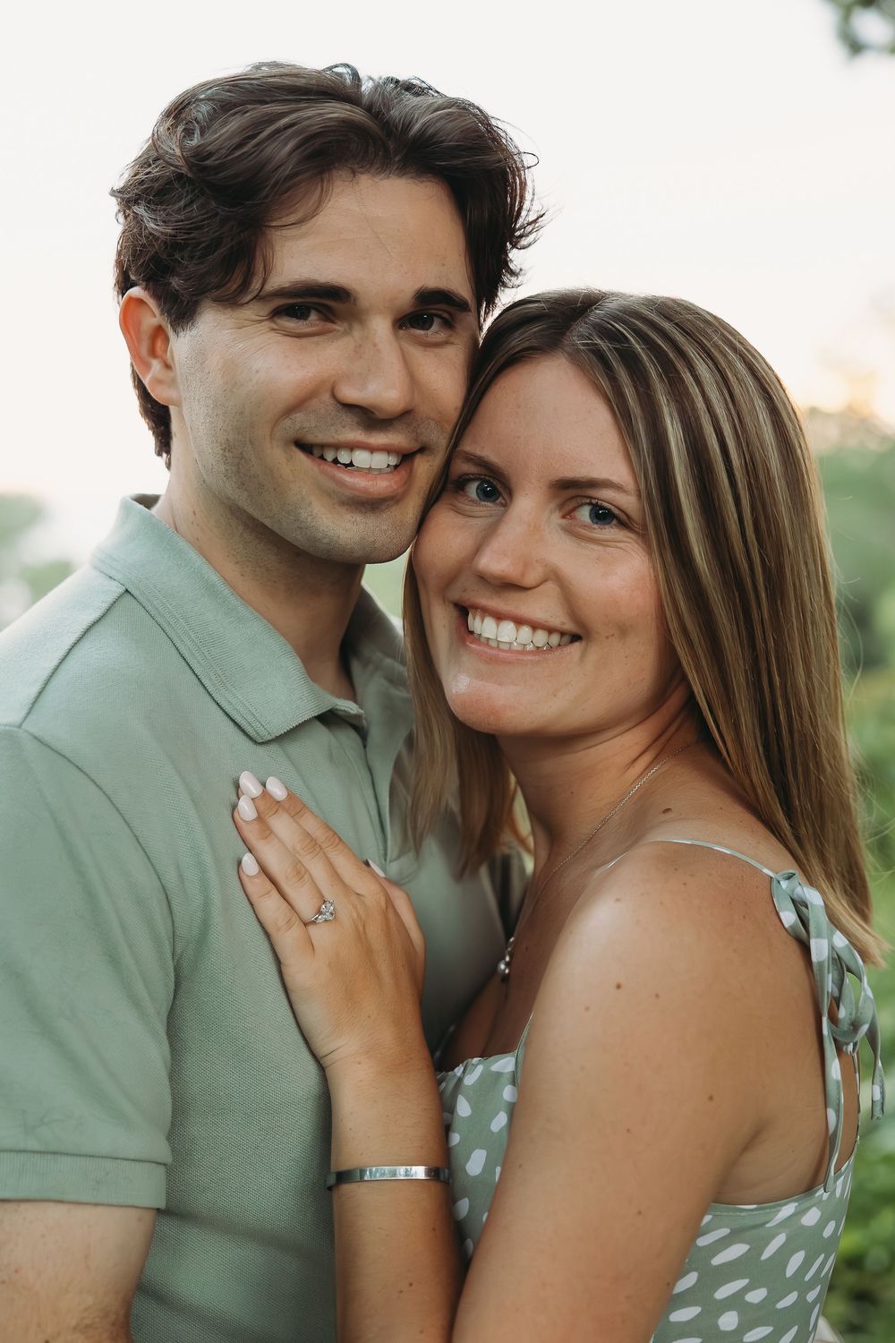 Smiling couple posing cheek to cheek outdoors, woman wearing engagement ring and polka dot dress, man in green polo shirt.