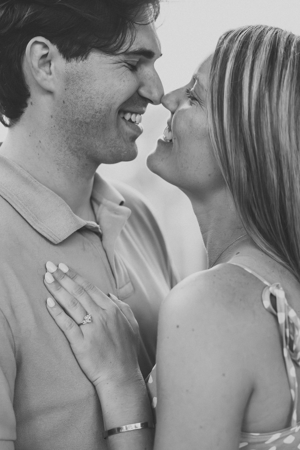 Black and white photo of a smiling couple touching noses, woman showing engagement ring on mans chest.