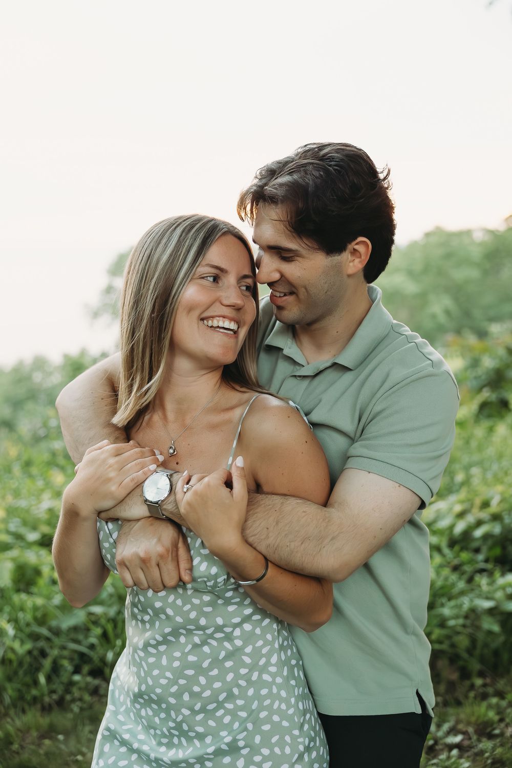 A smiling couple embraces outdoors, woman in floral dress and man in green polo, surrounded by lush greenery.