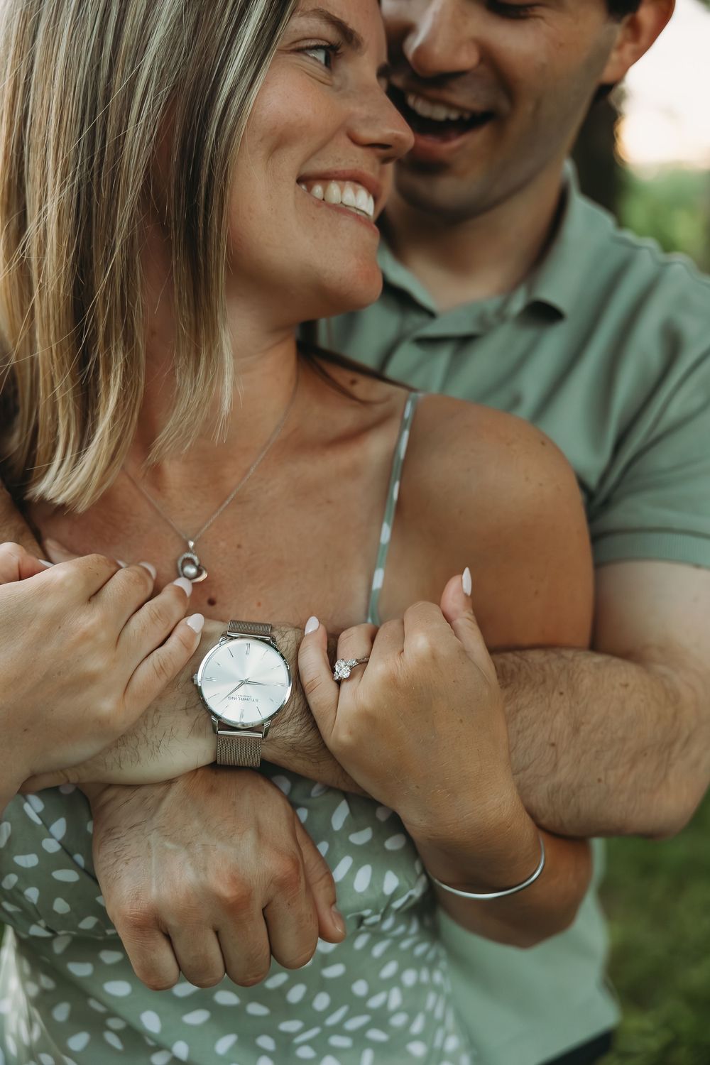 Smiling couple embracing outdoors, woman displaying engagement ring and watch while man hugs her from behind.