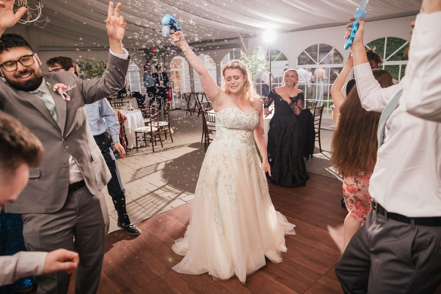 Bride dancing alone with her eyes closed, holding a bubble gun overhead as bubbles fill the reception tent around her.