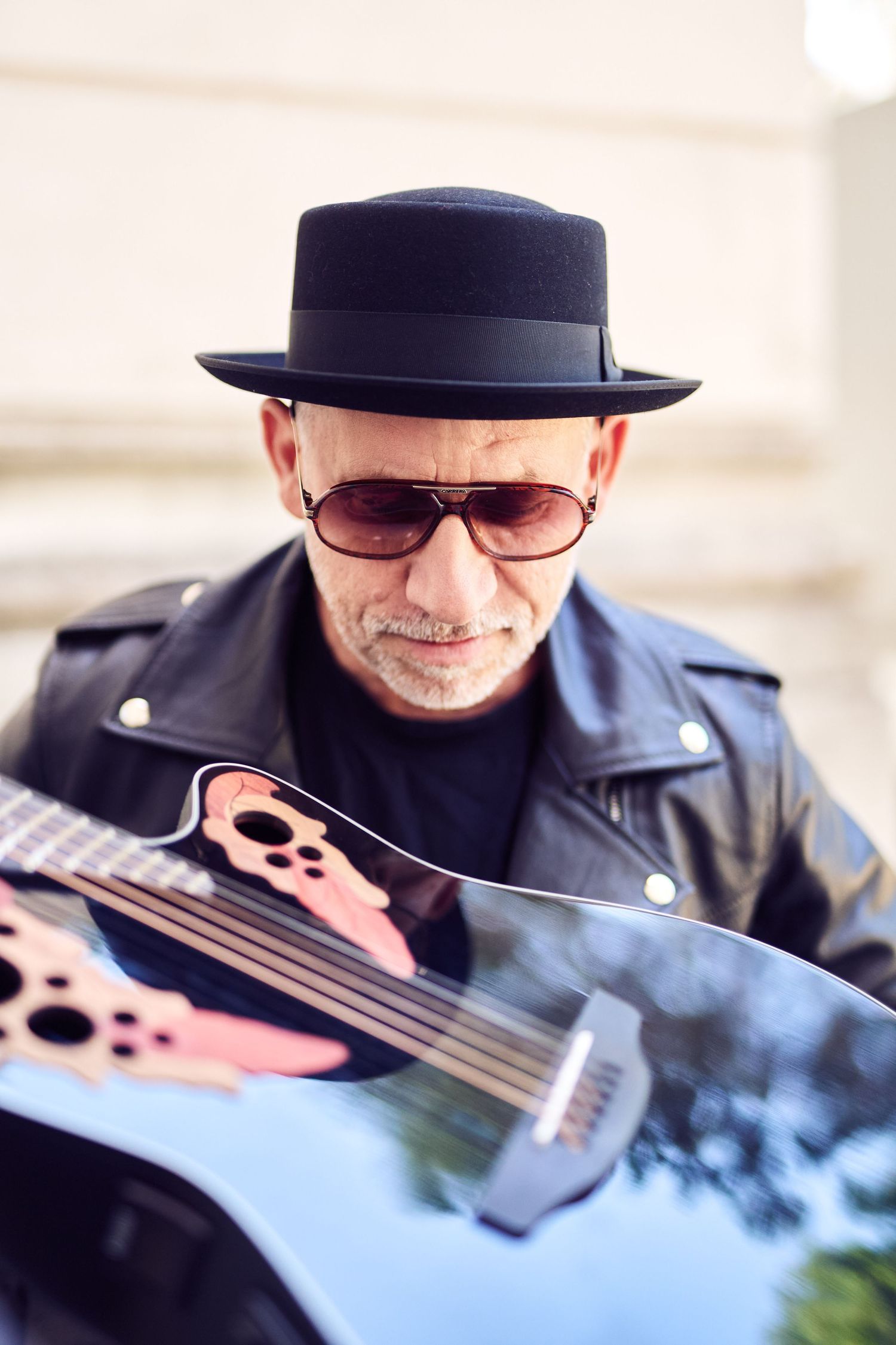 Musiker in schwarzer Lederjacke, Mütze und Sonnenbrille, die im Freien eine schwarze Akustikgitarre spielt.