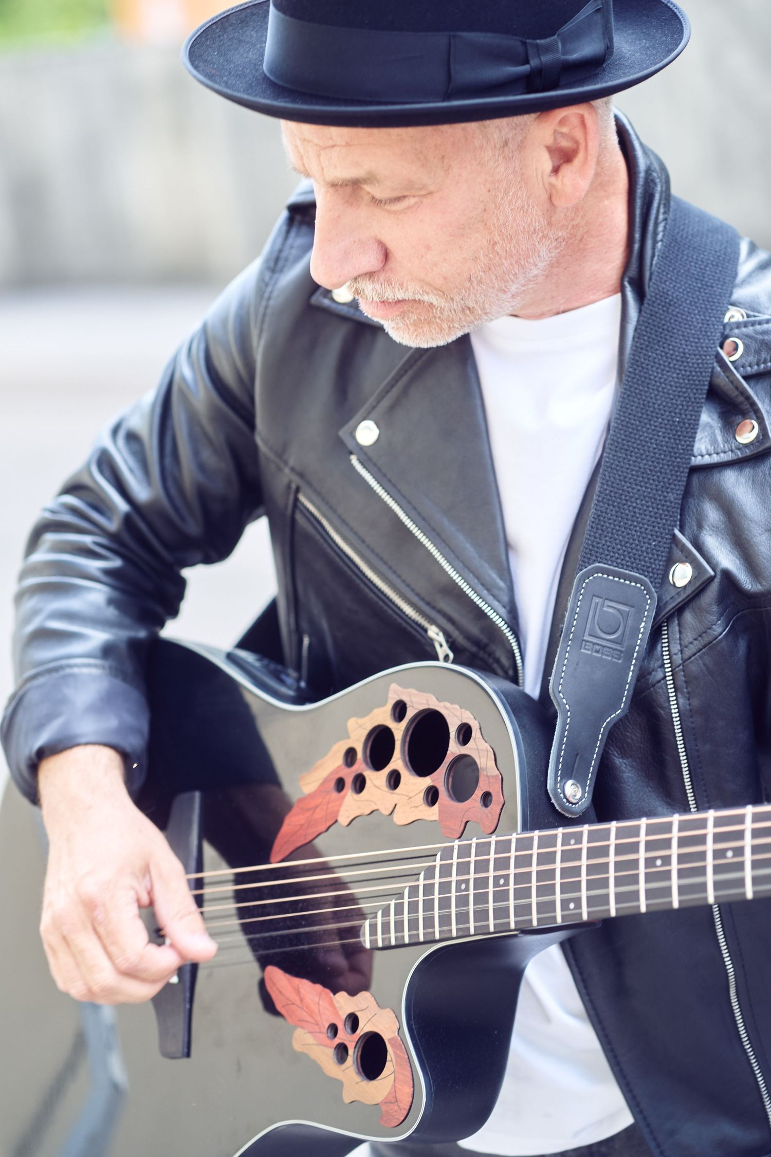 Musiker in schwarzer Lederjacke und Hut, der im Freien akustische Gitarre spielt.