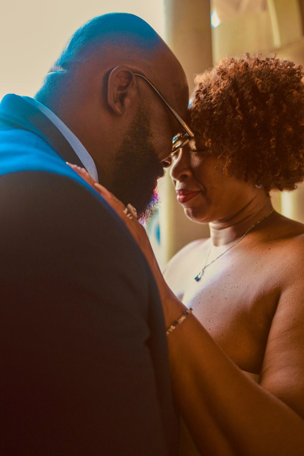 A couple shares an intimate moment, foreheads touching, dressed elegantly in warm golden light.