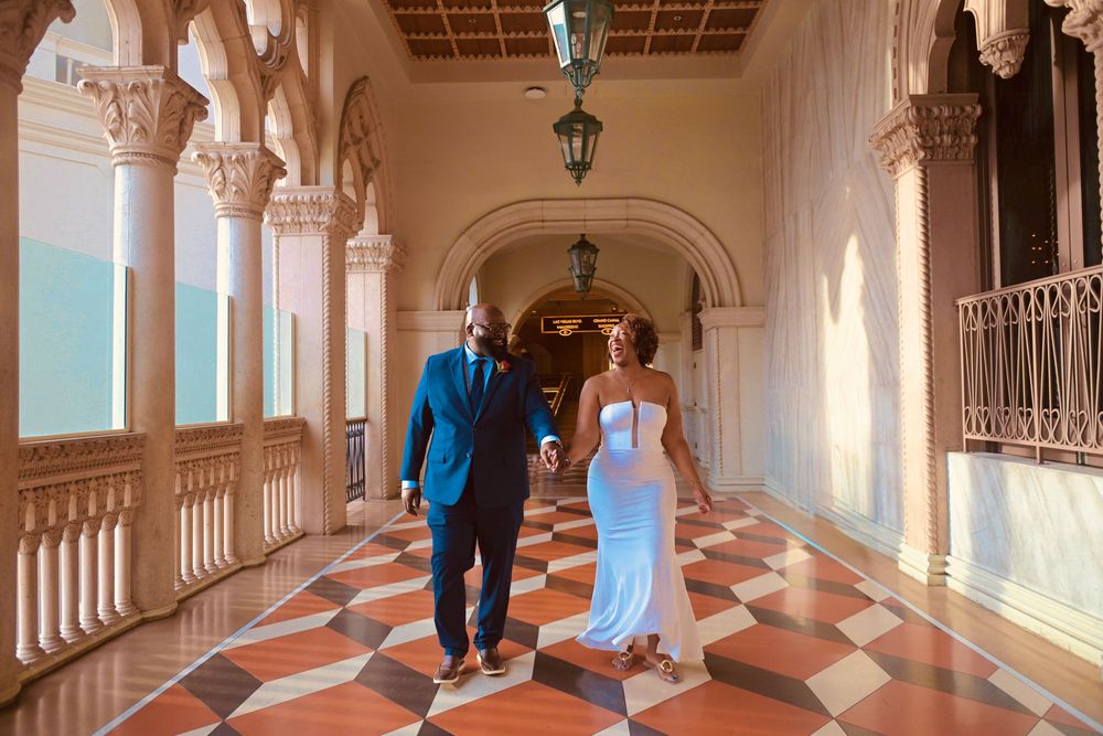 Couple walks hand-in-hand through elegant Venetian-style corridor with ornate columns and checkered marble floors.