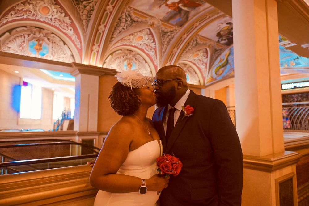 Newlywed couple sharing a kiss in an ornate wedding venue with colorful frescoed ceilings and warm lighting.