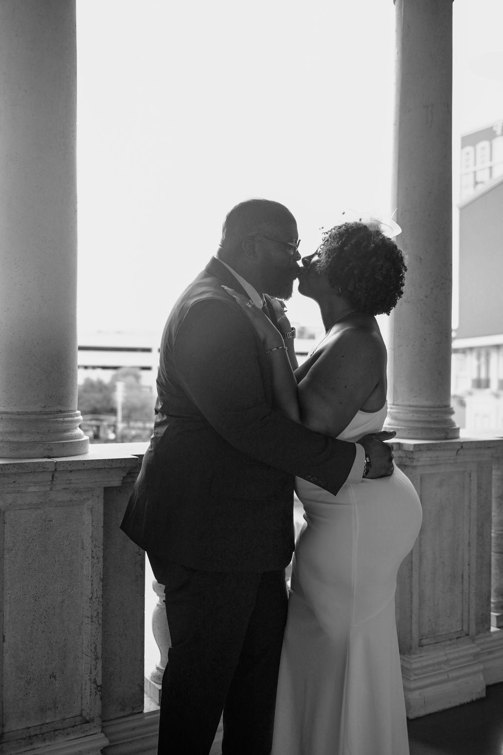 Black and white photo of a couple kissing on a columned balcony in wedding attire.