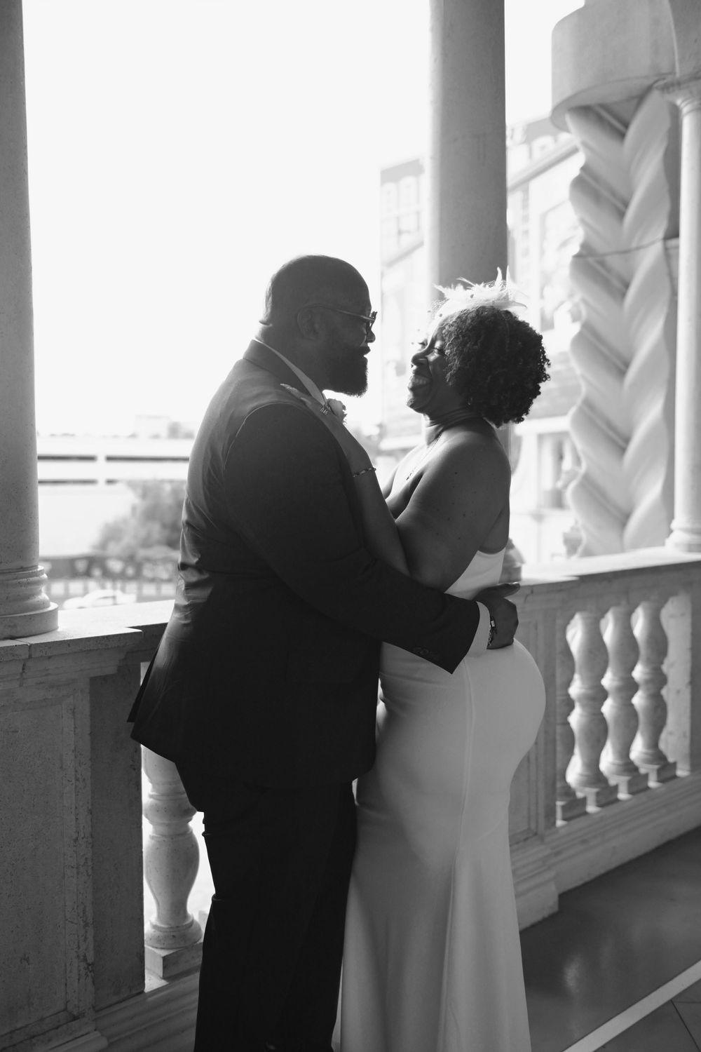 Black and white photo of a couple embracing on an elegant balcony, the bride in a white gown and groom in a suit.