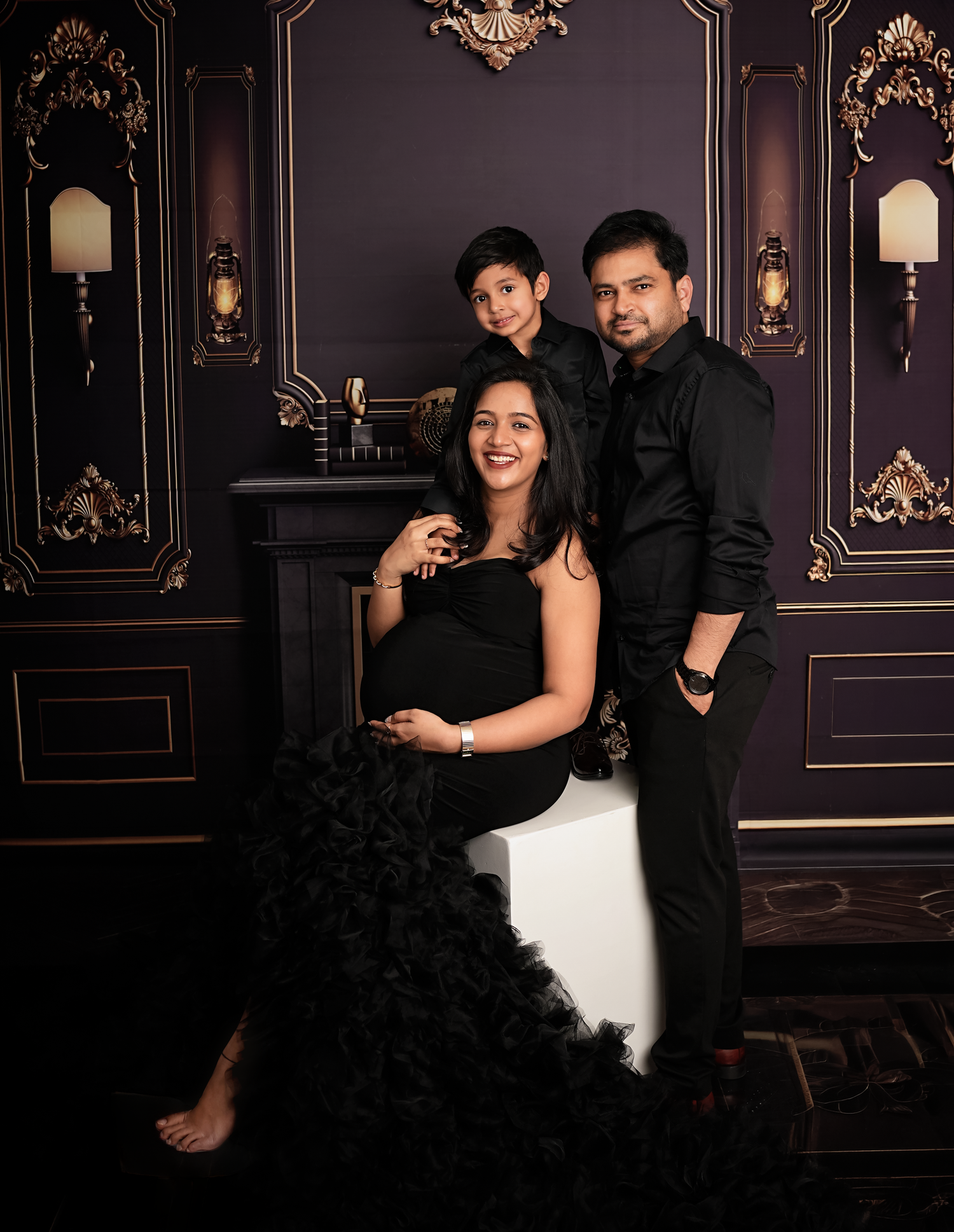 Elegant maternity photo of a pregnant woman in black dress with husband and son against dark ornate backdrop.