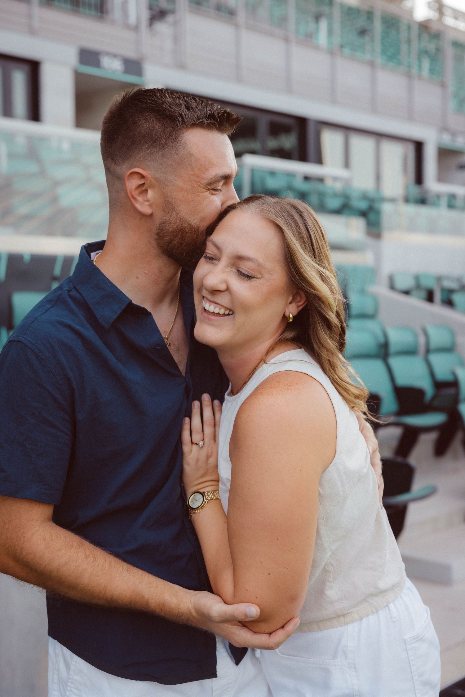 A woman smiles and leans against her fiancé's chest as he kisses her on the head following their surprise proposal at CPKC stadium in KC.