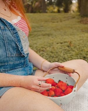 Red-haired woman in denim overalls sitting outdoors holding a white colander filled with fresh red strawberries.