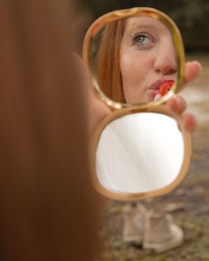 A young woman with red lips checks her makeup in a small gold compact mirror outdoors.