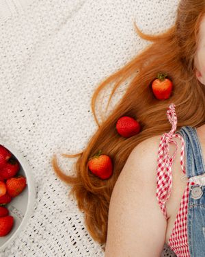 Red-haired girl lying on white knit blanket with fresh strawberries placed in her flowing auburn hair.