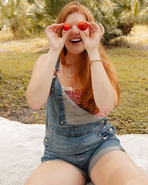 A cheerful redhead girl in denim overalls holds strawberries over her eyes while sitting outdoors on a white blanket.
