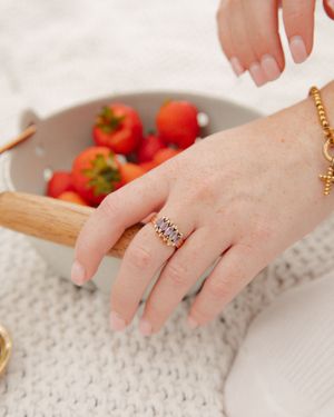 A hand wearing a gold gemstone ring and bracelet rests on a knit surface beside a bowl of fresh strawberries.