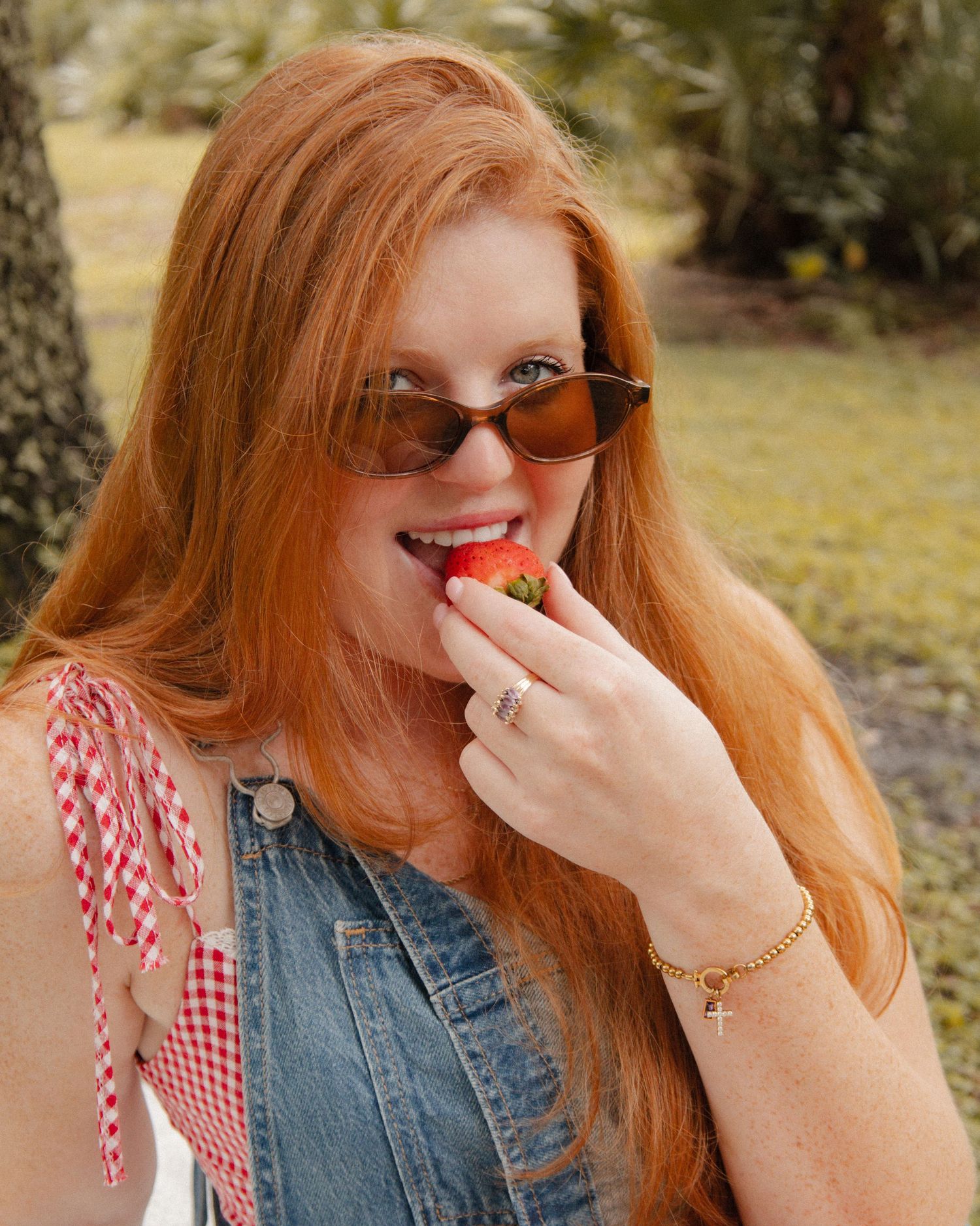 Red-haired woman in sunglasses and denim vest eating a fresh strawberry outdoors near trees.