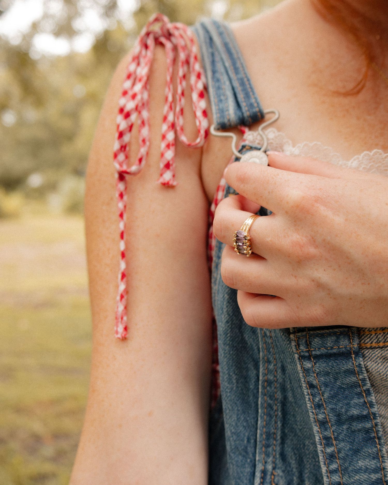 Woman wearing denim overalls with red gingham tie detail and gold rings, outdoors.