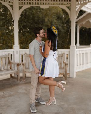 Couple sharing a kiss under a white gazebo, woman wearing graduation cap and white dress.