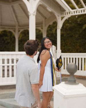 Smiling graduate in white dress holds cap and stole while holding hands with partner near gazebo.