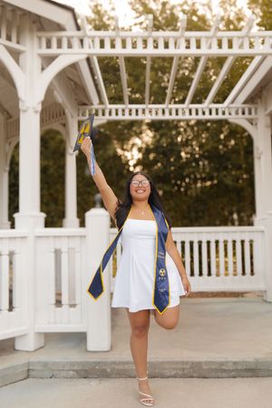 Joyful graduate in white dress celebrating under a white gazebo with diploma raised.