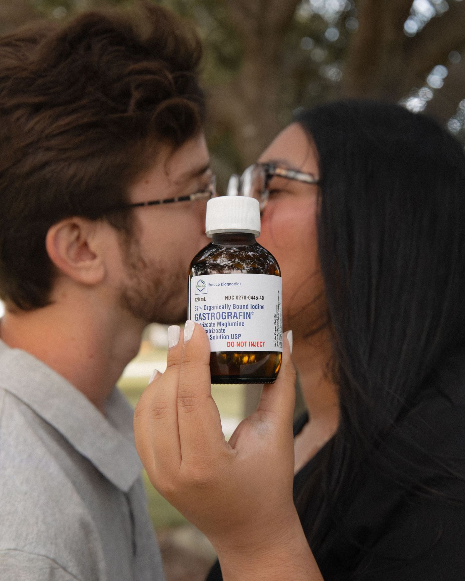 Couple kissing behind a medicine bottle held up to the camera outdoors.