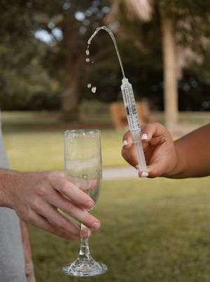 Hand using a medical syringe to pour liquid into a champagne flute outdoors.