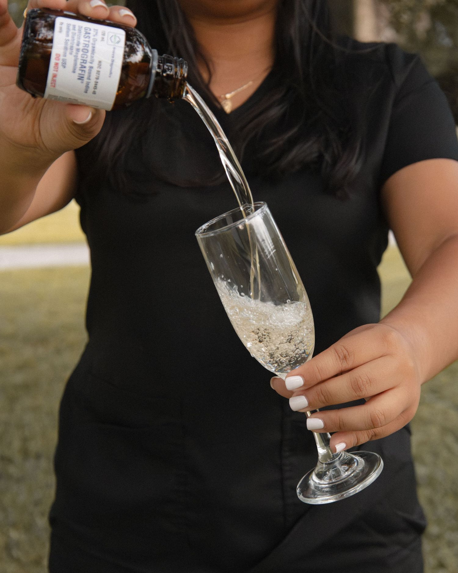 Woman in black scrubs pouring champagne from a medicine bottle into a flute.