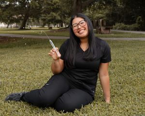 Smiling woman in black scrubs sits on grass holding a syringe outdoors.