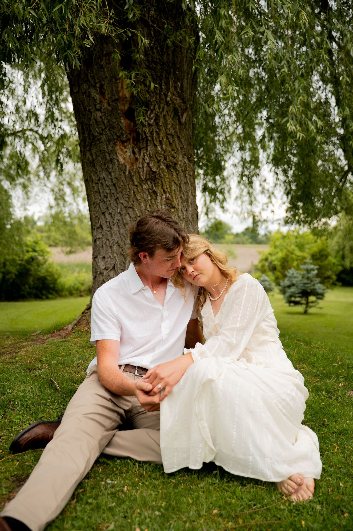 A couple in white attire sits romantically under a large willow tree on green grass in a serene outdoor setting.
