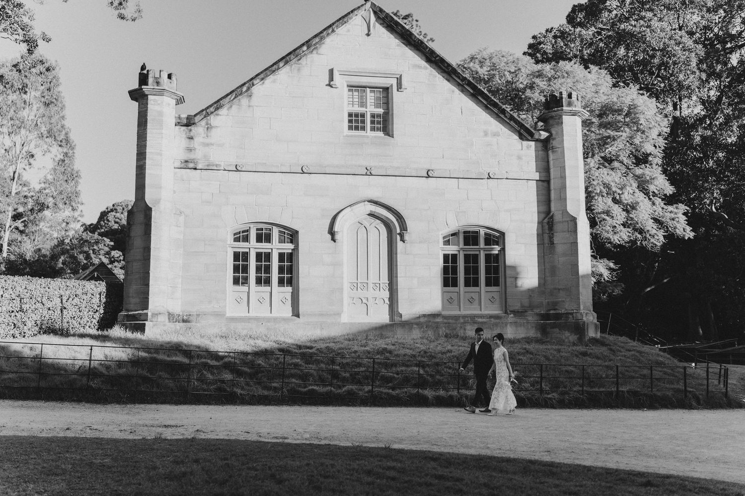 Black and white wedding photo of couple standing before a historic stone building surrounded by trees.
