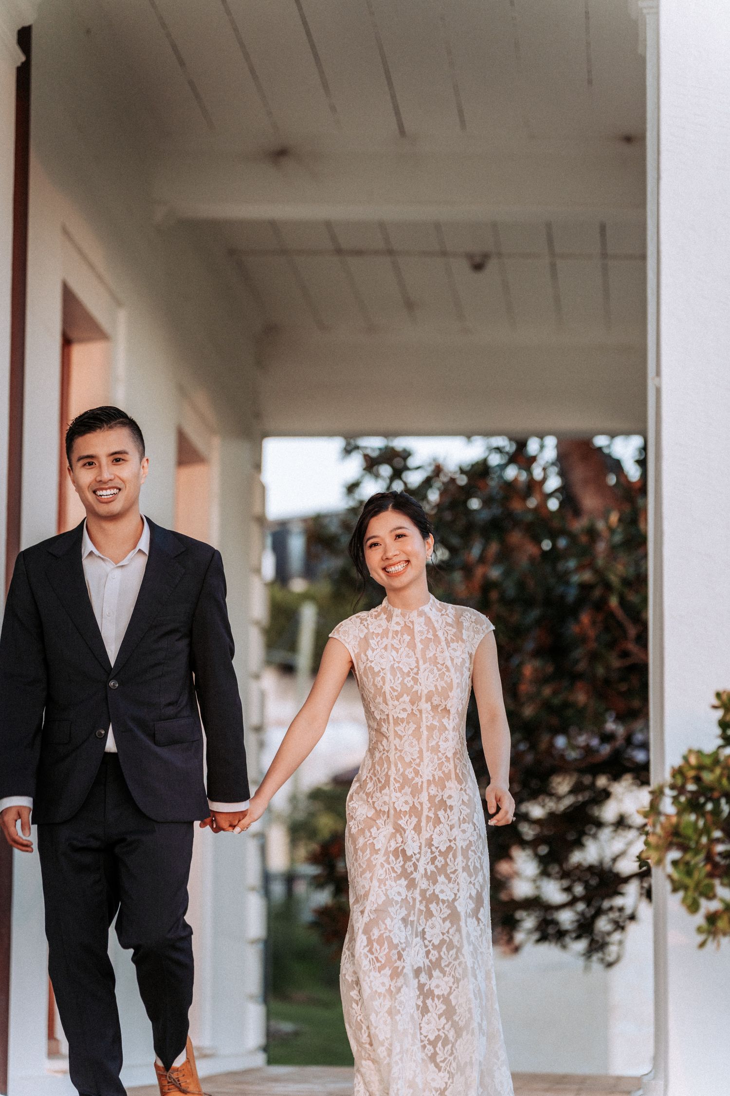 Smiling couple holding hands, woman in white lace dress and man in black suit, walking on a porch.