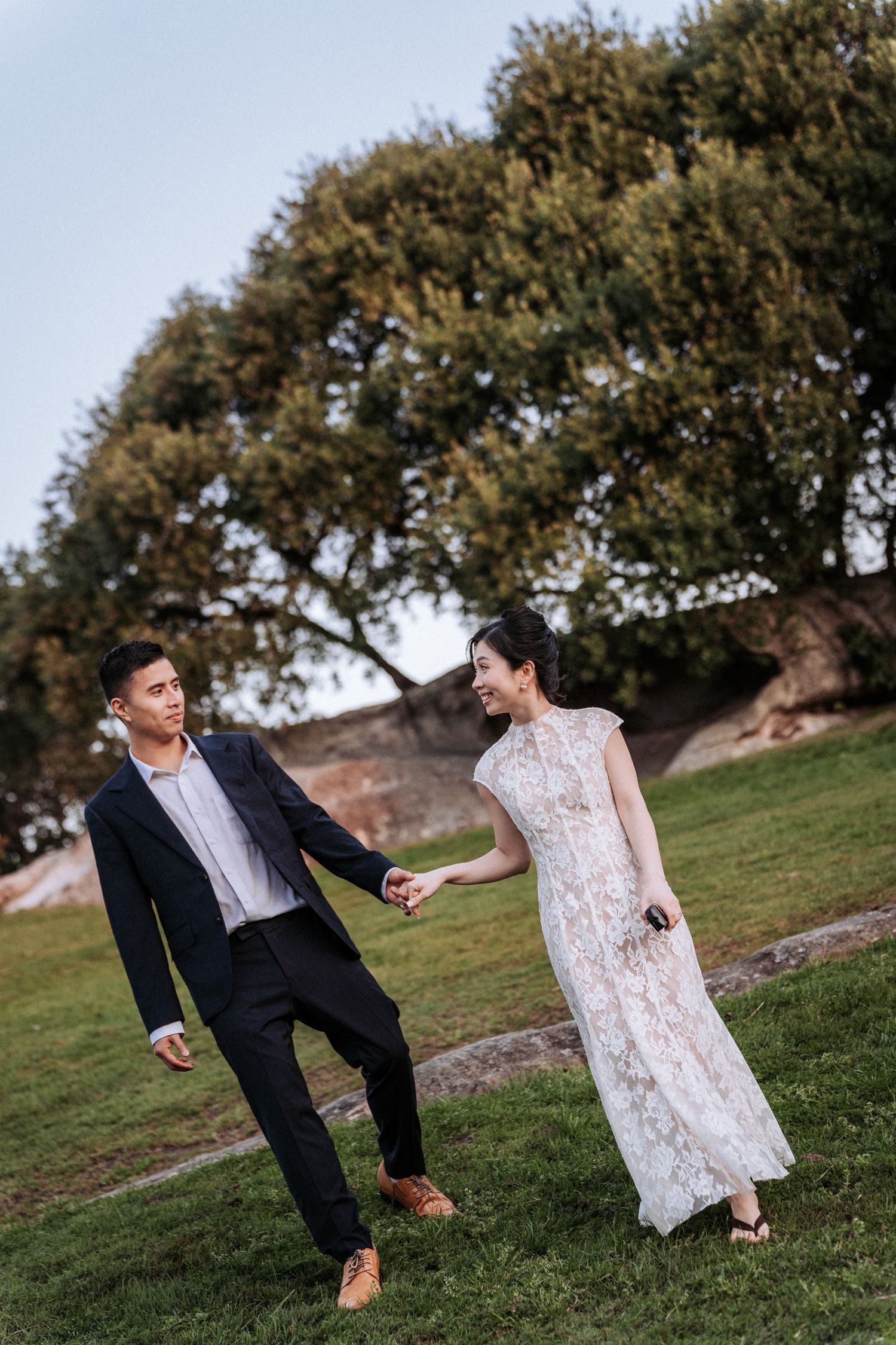 Couple in formal wear holding hands and walking on green grass with a large tree in the background.