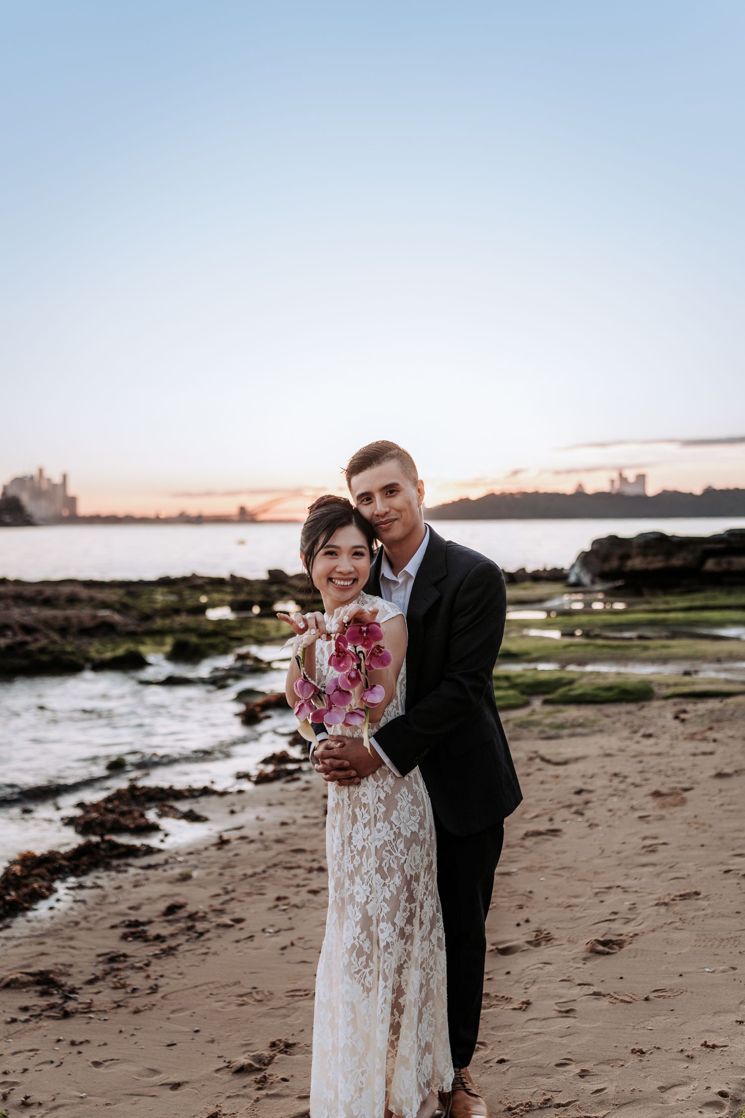 Couple embracing on beach at sunset, bride in lace gown holding pink bouquet, groom in black suit, harbor backdrop.