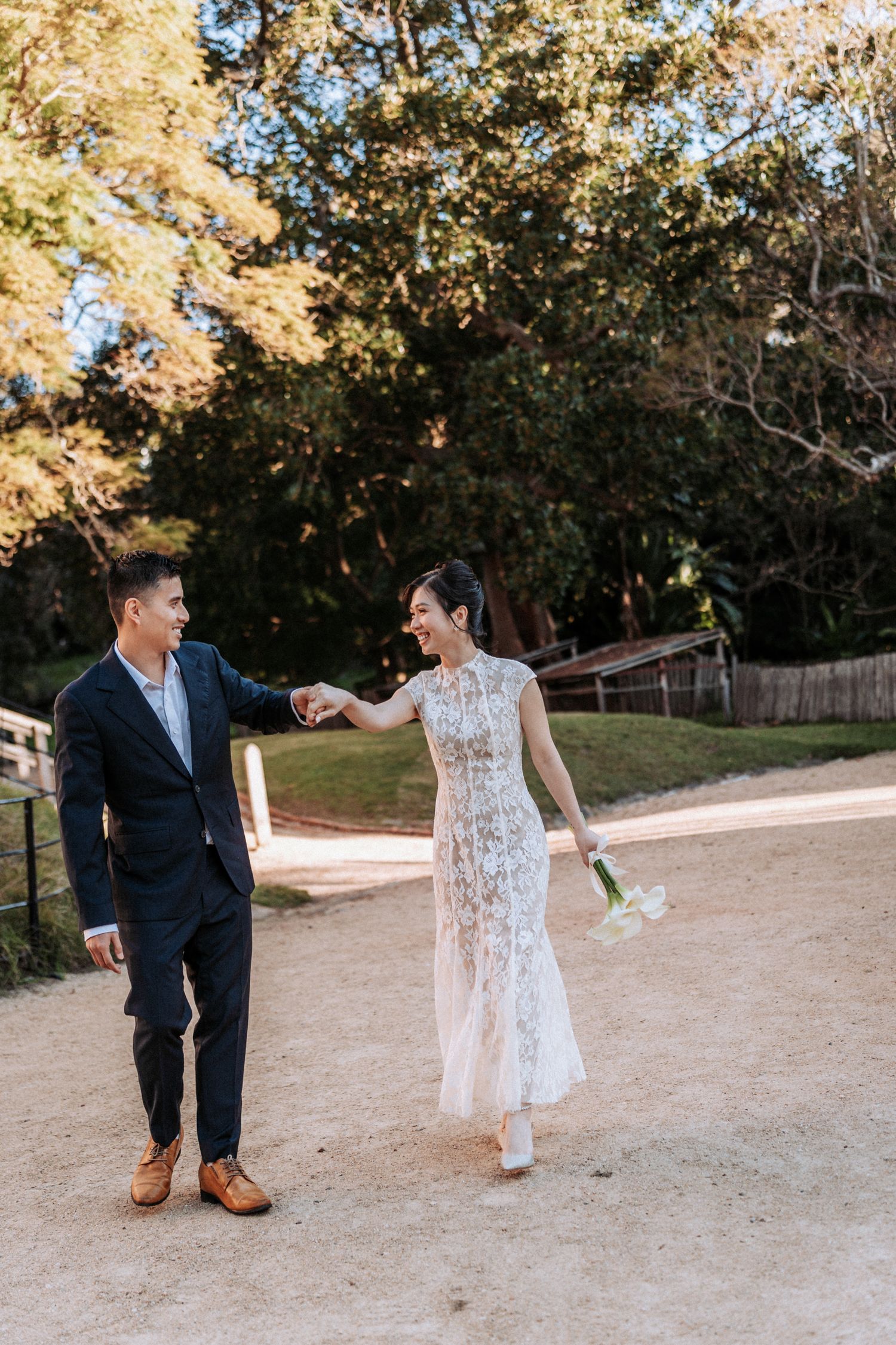 A bride and groom hold hands while walking outdoors on a sandy path surrounded by lush green trees on their wedding day.