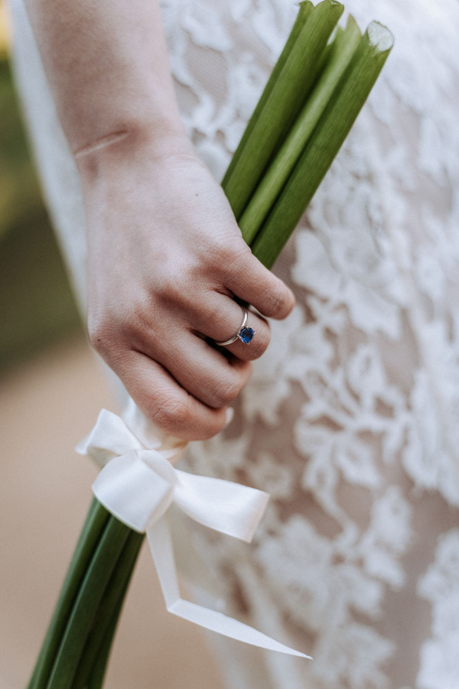 Bride holding green stems tied with white ribbon, wearing a blue sapphire engagement ring, in a white lace wedding dress.