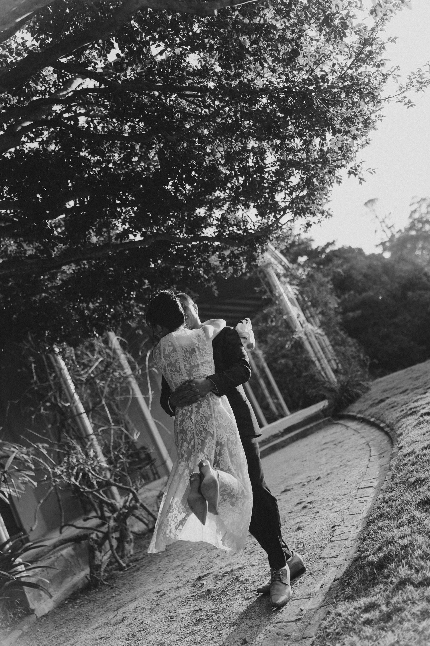 Black and white photo of a couple embracing on a garden path, bride in lace gown and groom in suit, surrounded by lush trees.
