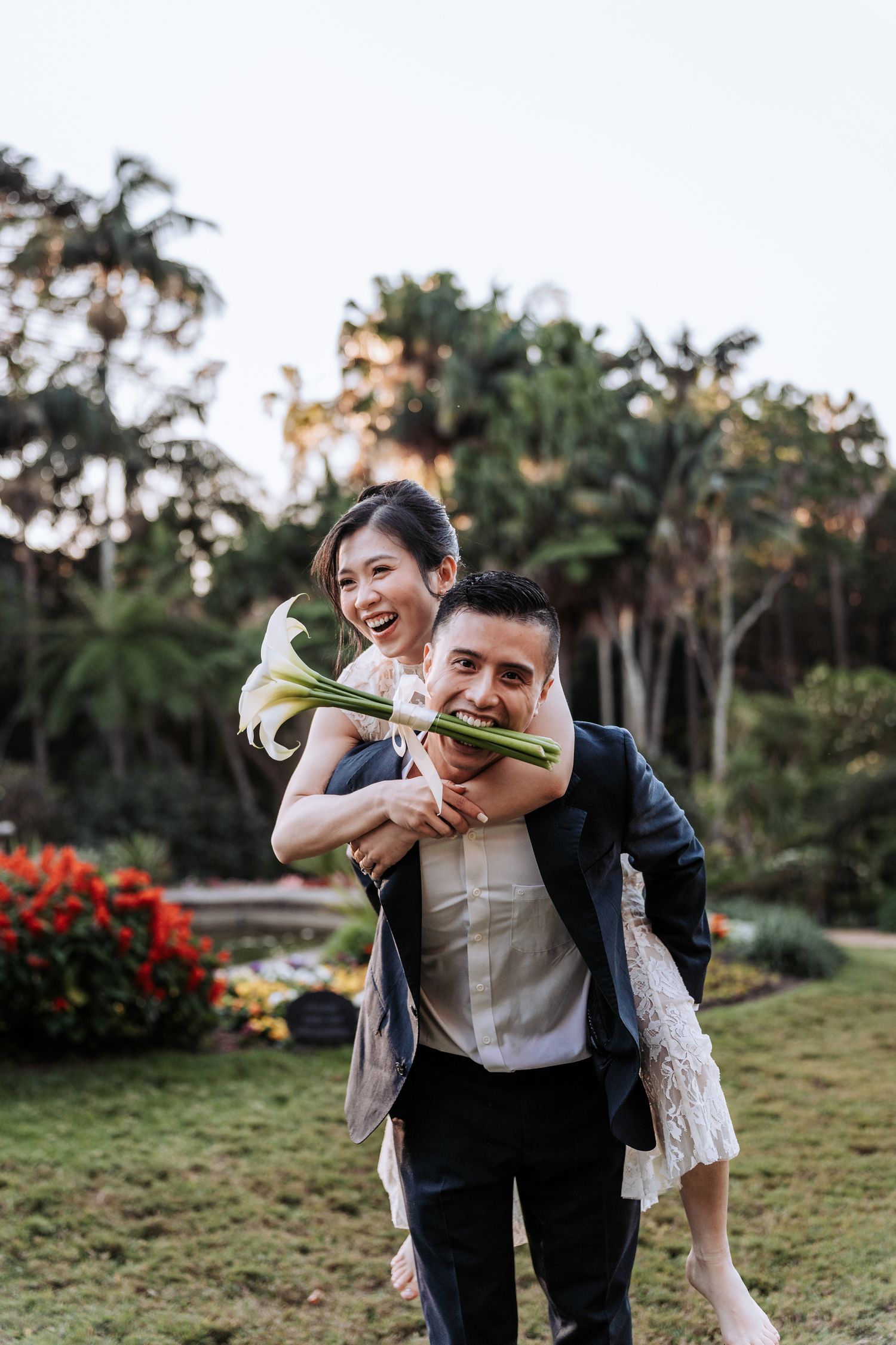 A joyful couple in a lush garden, man giving woman a piggyback ride, she holds white lilies, both smiling, dressed for wedding.