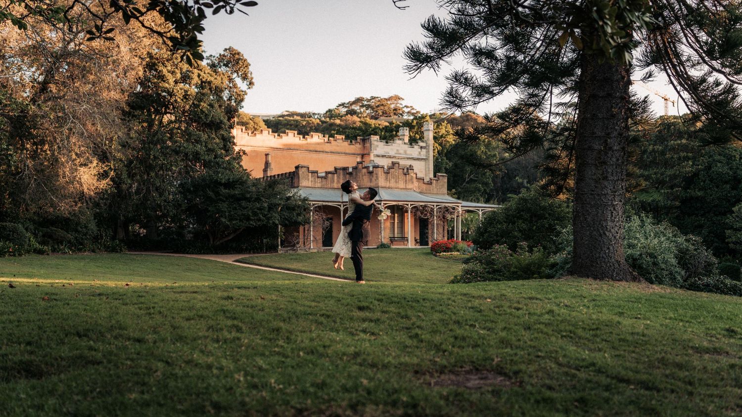 Couple dancing on lush green lawn beside historic manor house surrounded by tall trees at golden hour.