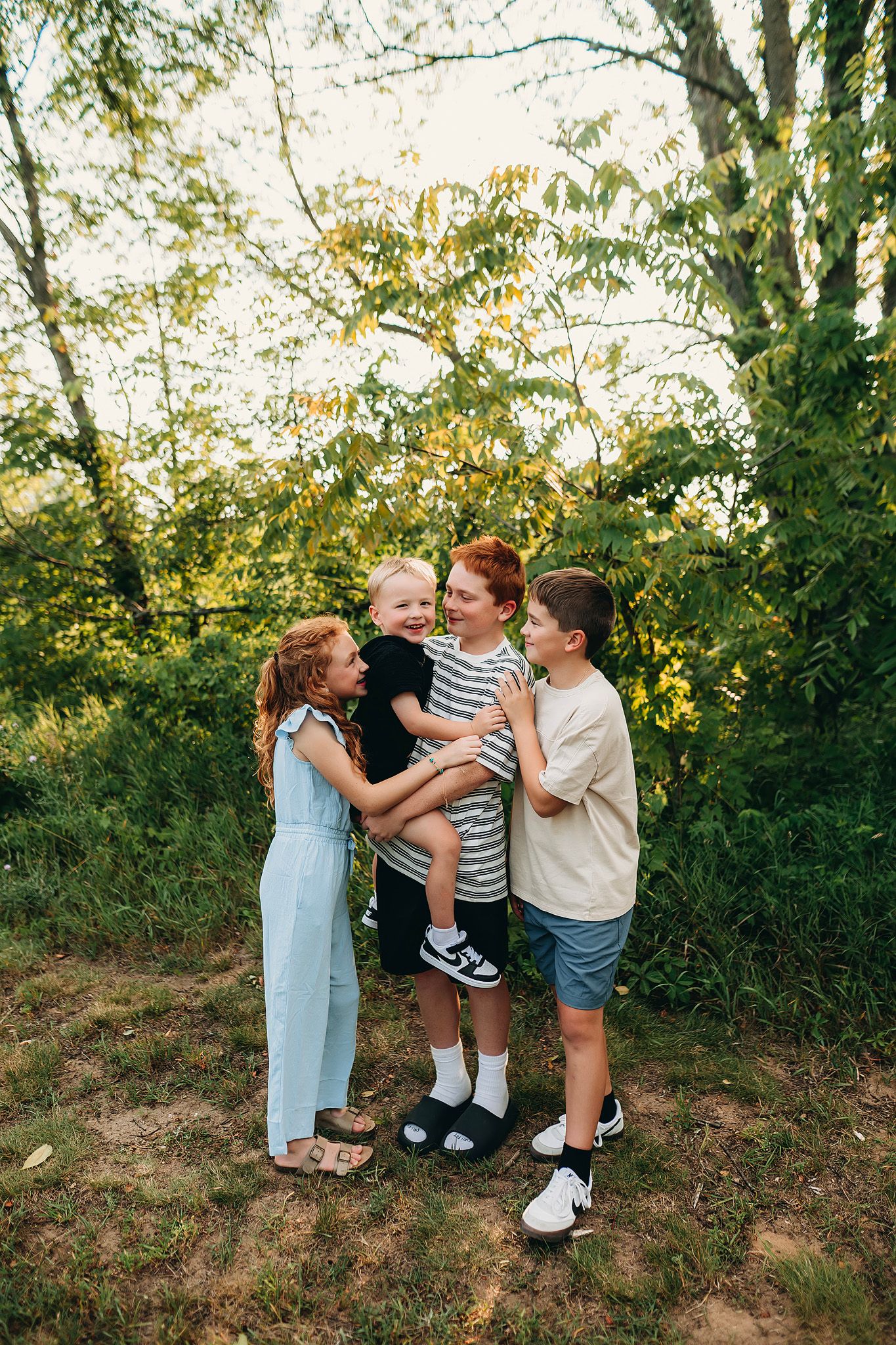 Four children laughing and hugging outdoors in a lush green forest setting during golden hour.
