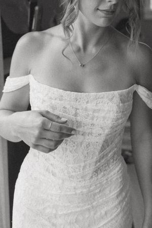 Black and white photo of a bride in an off-shoulder lace wedding dress adjusting her gown, wearing a delicate necklace.