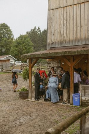 Guests shelter under a rustic wooden barn overhang during a rainy outdoor wedding at a farm venue.